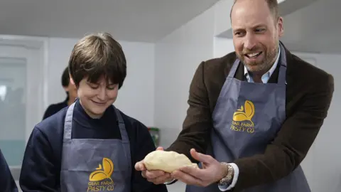 Prince William is holding a pasty. He is wearing a brown jacket and a blue apron.