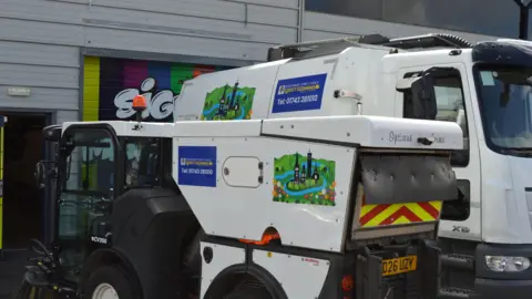 Shrewsbury Town Council Two white and black street cleaning vehicles are parked up outside a warehouse. A smaller one on the left hand side has text reading "Optimus Grime" on the back. 