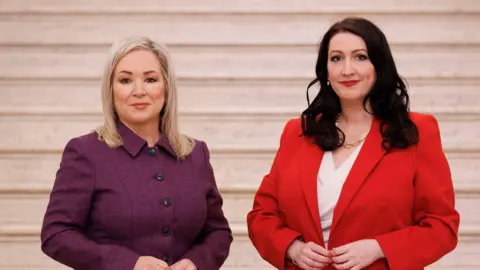 Michelle O'Neill is wearing a purple button up suit. She has blond hair to her shoulders and her arms by her front. Emma LP is wearing a red blazer open above a white v neck shirt. She has a gold pearl necklace on. She has long curly brown hair. Her arms are by her front. Both women stand side by side in front of the Stormont Great Hall steps. 