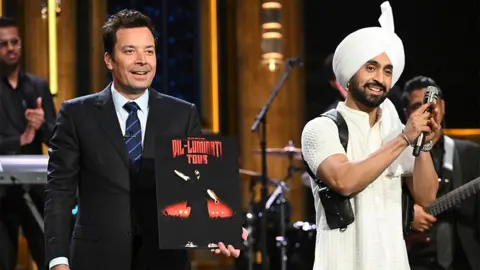 El presentador estadounidense Jimmy Fallon viste un traje negro y una camisa blanca con Diljit Dosanjh, quien viste de blanco con un micrófono negro en su mano, mientras hace un gesto de oración con sus manos hacia la audiencia.