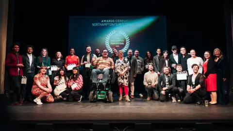 Khandie Photography A large group of people on a stage, in front of a screen. Some are kneeling, others are standing up. Some are holding clapper boards. They are all smiling and looking at the camera. 
