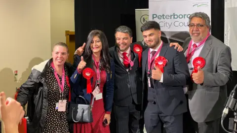 Ben Schofield/BBC Five people, wearing Labour rosettes pose for a picture. Four of them are giving a thumbs up sign. In the background are banners advertising Peterborough City Council. 