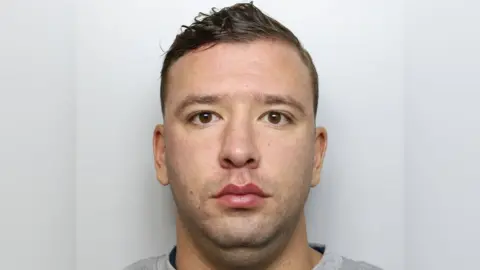 A custody image of a man with a round face, brown eyes and brown hair. He stares straight down the lens with a grey background behind him.