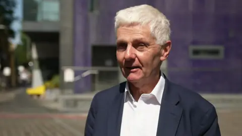 A man with white hair wearing a blue suit and white shirt is being interviewed outside a modern corporate building. 