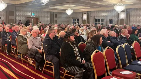 Hundreds of people are sitting in a room on red seats. There are a number of chandeliers hanging from the ceiling and five vacant seats in the front row of the audience. 