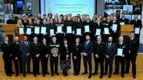 Over 50 members of Lancashire Police staff stand in three rows, with some holding certificates and shields. Some are in uniform. A screen behind them says 'congratulations and thank you to all recipients for your service and dedication'.