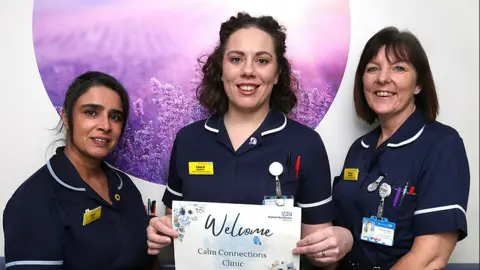 Three nurses are in uniform, with one of them, Laura, holding a welcome poster for the clinic.