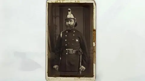 LFB A man stood upright in uniform with helmet on his head. The image is small and black and white.
