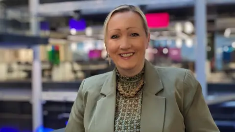 Rebecca Evans photographed at BBC Wales headquarters with coloured panels behind as she stands by a railing. She wears a khaki suit and patterned white and khaki blouse with a large necklace and has blonde hair tied back.