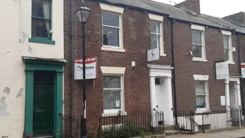 The outside of the terraced building which has a white door, one window on the ground floor and two on the first floor. It has a orange and blue logo on the windows and white sign above the door. The building on the left is cream with peeling paint. The building on the right has a white sign outside of it.