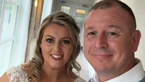 Samantha Jones and Trevor Jones smiling into the camera. Samantha is standing on the left and has blonde, curled hair and is wearing dark pink lipstick. Trevor is standing closer to the camera and has short, dark hair and is wearing a white shirt.
