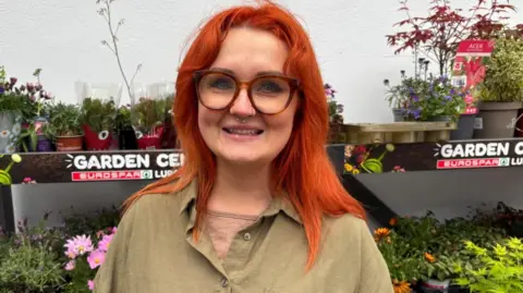 A woman with bright orange hair is smiling into the camera. She has a nose ring and is wearing large, brown glasses, a green short sleeved shirt and a green t-shirt. She is standing in front of a stall of flowers in plastic pots.