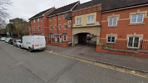 A modern, brick-built three-storey building is on the left of the photo, with a two-storey building on the right and an archway between them. Five vehicles are parked on the left hand side of the image.