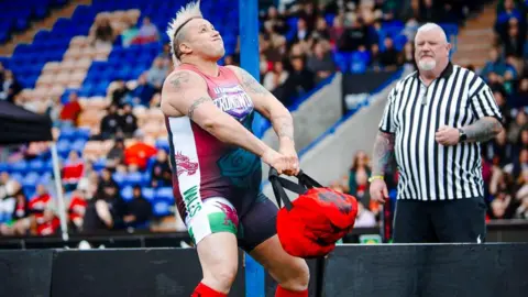 Harry Collins Sam straining to lift a an organ bag, with a referee and the crowd looking on