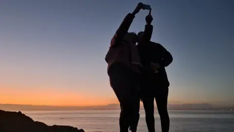 Naomi Dring A silhouette of three women is seen posing for a selfie on Somerset beach in Clevedon. The setting sun behind them casts a light orange glow over the Severn Estuary