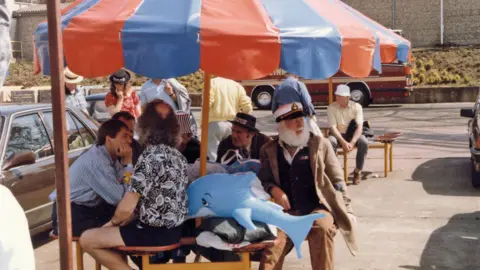 Only Fools and Horses Appreciation Society People sitting around a seaside table under a striped umbrella, including Uncle Albert, with a large inflatable dolphin, parked cars and a coach in the background.