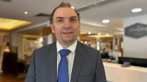 Simon Thake/BBC A man with thinning hair stands in a hotel lobby in a grey suit and blue tie.