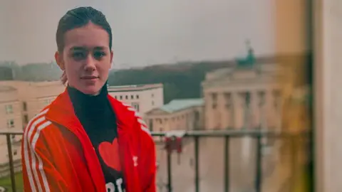 Brunette white teenage girl standing on a balcony. Background is blurred buildings. She is wearing a red sports jacket and black polo neck top.