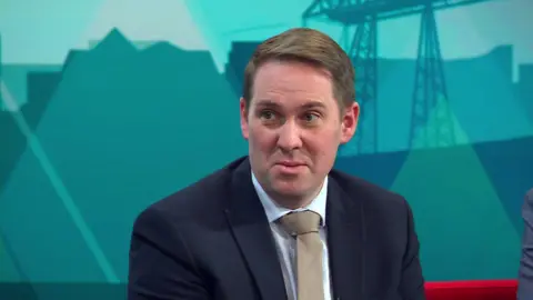 Labour MP Sam Rushworth. He is wearing a navy suit and beige tie. He has short brown hair. He is sitting on the red couch on the set of BBC Politics North.