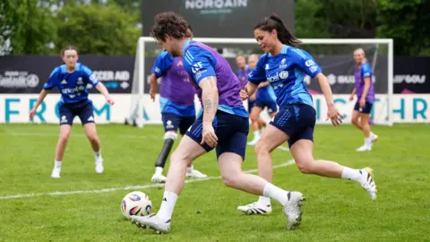 A man dressed in a blue, purple and black football kit, dribbles with a ball past a woman wearing a similar kit. Other players are watching on in the background with a goal visible behind them.
