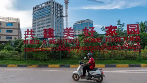 Jonathan Head/ BBC A man riding past a red sign saying 'Let's build a beautiful home together'  