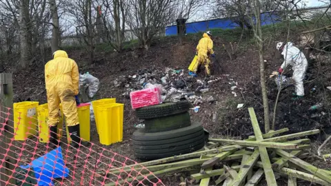 Workers wearing either white or yellow jumpsuits work to clear mess dumped on a muddy verge, behind which is the A14. The mess includes wooden beams, general litter and tyres. A series of yellow bins are being used to store some of the rubbish.