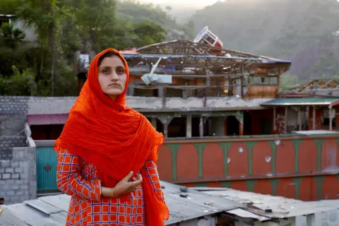 TASEER BEYG / BBC Nimra, 16, wearing an orange headscarf and a patterned outfit stands in front of the damaged mosque, mountains visible in the distance.
