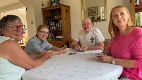 BBC Four people playing cards at a round table with a table cloth on. Left to right - An older woman in a light green shirt she is next to a teenage girl wearing a stripy top, next to her is a elderly bald man in a grey T-shirt and a middle aged woman with ginger hair and a pink top.