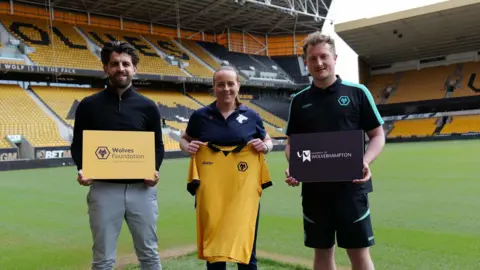 Two men are holding signs, one which says "Wolves Foundation" in black with an orange background and the sign on the right says "University of Wolverhampton". In the centre a woman holds up an orange Wolverhampton Wanderers Football Club shirt. 