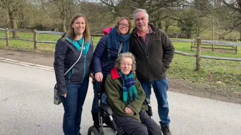 Catie Alison is in a wheelchair outside surrounded by her two daughters and husband