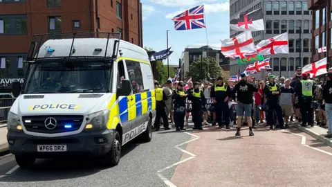 A police riot van parked on a bridge, which is blocked by protesters standing behind a line of police officers. The group hold flags, including Union and St Gorge flags and the Welsh flag. 