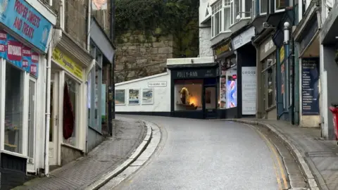 BBC The narrow lane leads up hill and turns sharply to the left. There are small shops lining it and no people are in sight.