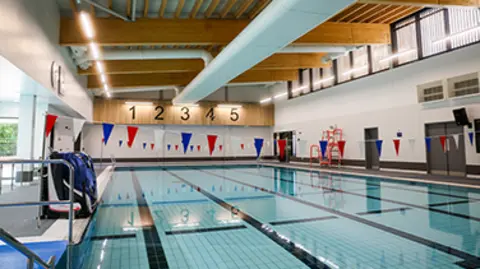 Everyone Active A swimming pool with flags going across it and the numbers 1 to 5 on a wooden board in the background. 
