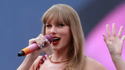 Taylor Swift waves as she sings into a glittering pink and orange microphone on stage at a concert. She has shoulder-length blonde hair and wears a necklace and red strapped outfit.