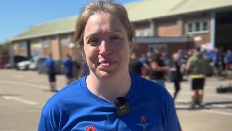 Lauren Carter/BBC A woman in a blue T-shirt smiling at the camera. There are crowds behind her out of focus.