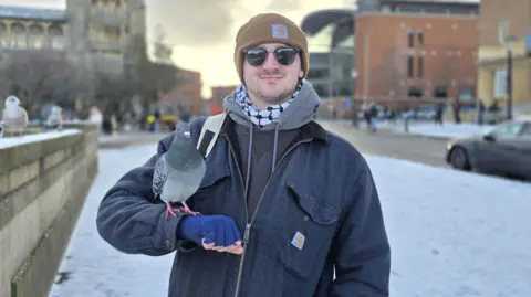 Paul Moseley/BBC Niall Adams is wearing a woolly beanie hat, sunglasses and several layers of clothes, including a blue jacket and a grey hoodie. A pigeon is perched on his right arm as he stands in a snow-covered street.