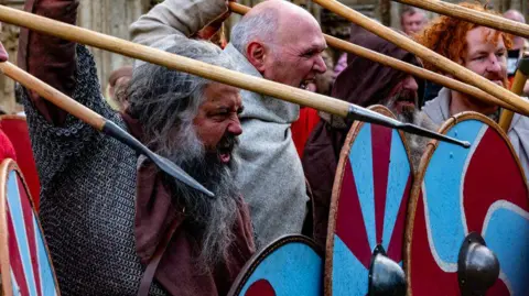 A group of men dressed as Vikings with angry expressions on their faces hold spears and shields as they look to charge at the enemy out of shot. 