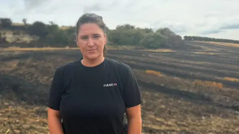 BBC/Jim Scott Farmer Tania Tallon is wearing a black t-shirt and has her dark, what looks like shoulder-length, hair tied back. She is standing on a field of what has been a crop such wheat or barley but which is now black and charred. There are low trees and bushes beyond and the sky is cloudy.