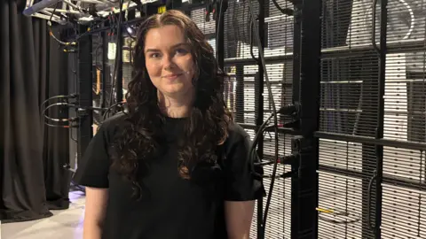 Johnny Reed/BBC Jasmine James, with long curly hair and a black T-shirt, stands against an indoor metal fence with wires in the background.