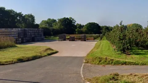 A short driveway on farm land, with grass verges, hedges, hay bales in the distance and concrete blocks on the left hand side of the image.