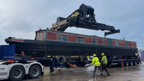 A canal boat is being lifted on to a haulage vehicle. Two men in high-vis clothing are pulling on ropes