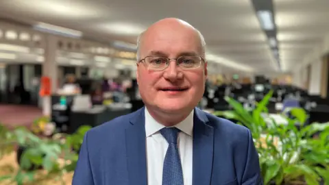 Council leader Simon Geraghty stands in the council's Wildwood offices. He has a bald top to his head with short white hair at the sides and wears half-rim glasses. He has on a blue suit, a white shirt and a blue tie with green plants behind him.