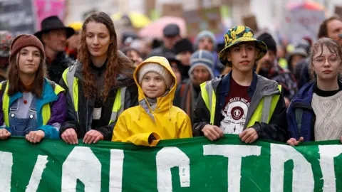 Swedish environmental activist Greta Thunberg photographed wearing a yellow coat, alongside four young demonstrators at a youth climate protest in Bristol. They are holding a green and white banner.