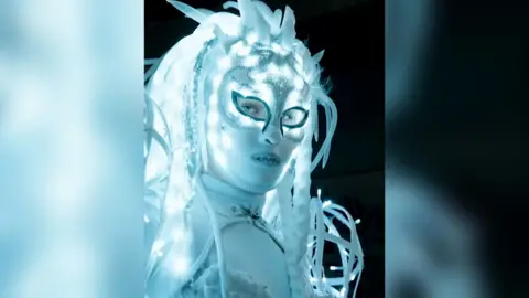 Performer in glowing white LED costume and sequined mask