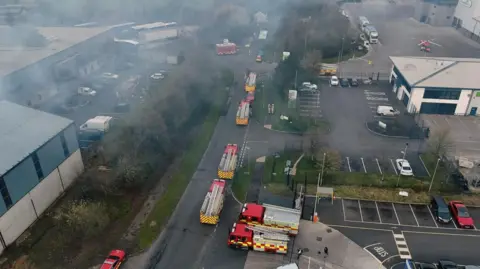 Jack McCausland A large number of fire service vehicles on a road beneath large plumes of smoke. The smoke is rising above the road and above trees and buildings.