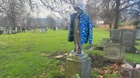 Friends of Anfield Cemetery The gravestone with Anfield Stadium in the background beyond an open grass area and other gravestones.