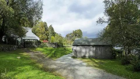 Brandlehow Cottage in the Lake District. The white-rendered cottage stands to the left at the end of a short gravel track. A wooden garage is to the right.