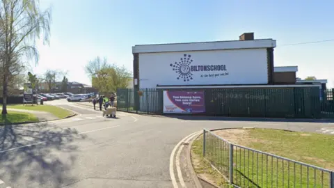 A building that is at least two storeys high with a white wall. On the building is a sign that reads Bilton School with purple squares in a spiral pattern. 