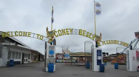 Entrance to the amusement park on a grey day with gates open and lettering above saying welcome to Coney Beach Pleasure Park. Attractions are visible behind including a caroussel.