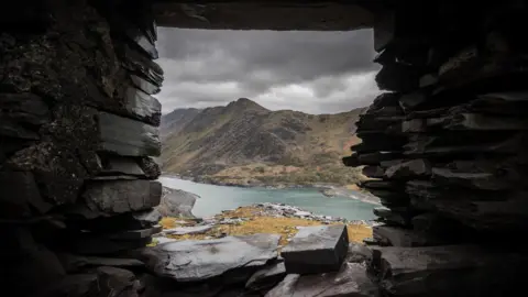 Getty Images is looking through a gray window without a glass in a list of a list. It introduces a view of the mountains in the Ereeri range, where it looks at the blue Llyn Peris lake, with the height of height in the back, the sky is dark with heavy black clouds, giving the entire image a sense of itself. It looks dark and dangerous.